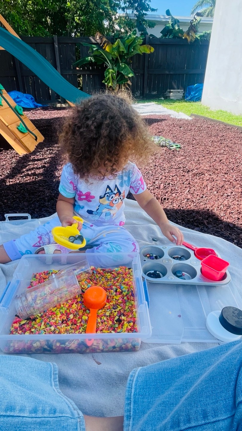 Child sitting outside using a colorful cereal sensory bin, scoops, and a muffin tin to sort objects by type.