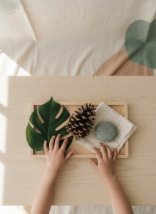 Overhead view of a preschool nature texture activity tray with a leaf, pine cone, large smooth stone, soft cloth, and exploring hands.
